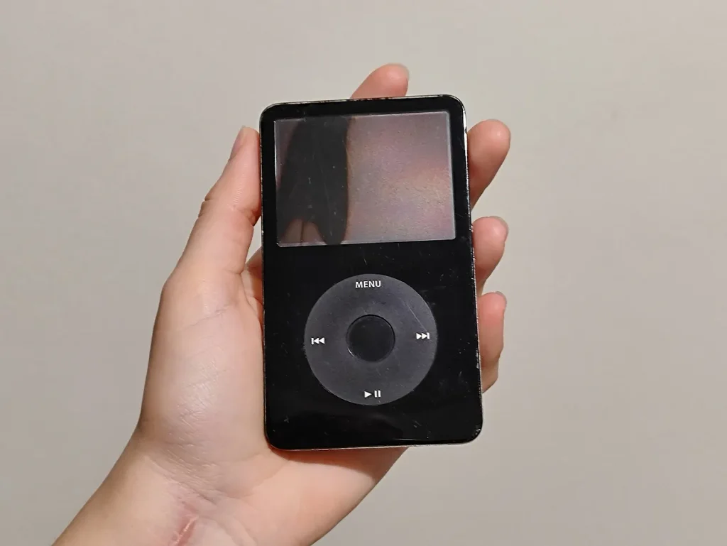 Mão branca segurando um iPod Video preto. O equipamento é um pouco maior que a palma da mão e em formato retangular, com uma tela em cima, ocupando metade do espaço, e na parte de baixo tem um círculo de controle de faixas e um botão circular no meio. Mão branca segurando um iPod Video preto. O equipamento é um pouco maior que a palma da mão e em formato retangular, com uma tela em cima, ocupando metade do espaço, e na parte de baixo tem um círculo de controle de faixas e um botão circular no meio.